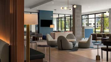 A modern hotel lobby with a seating area, a flat-screen TV, and large windows offering a view of the outside.