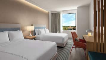 A hotel room with two beds, a desk, a chair, and a window with a view of houses.