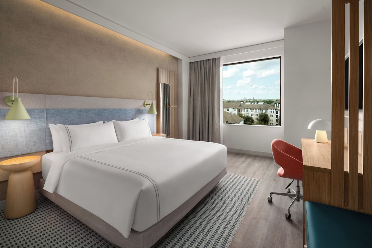 Una habitación de hotel moderna con una cama grande, lámparas de noche, un escritorio con silla y vistas a edificios a través del ventanal.
