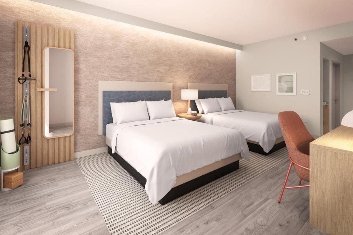 A modern hotel room with two beds, a wooden accent wall, a large mirror, and a desk with a chair.