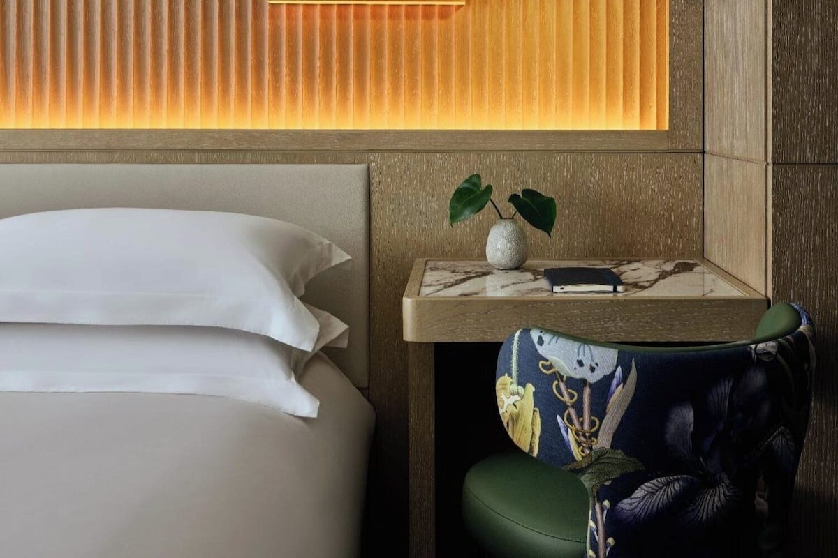 Una habitación de hotel con una cama, una mesita de noche, un florero con una planta y una silla de diseño colorido.