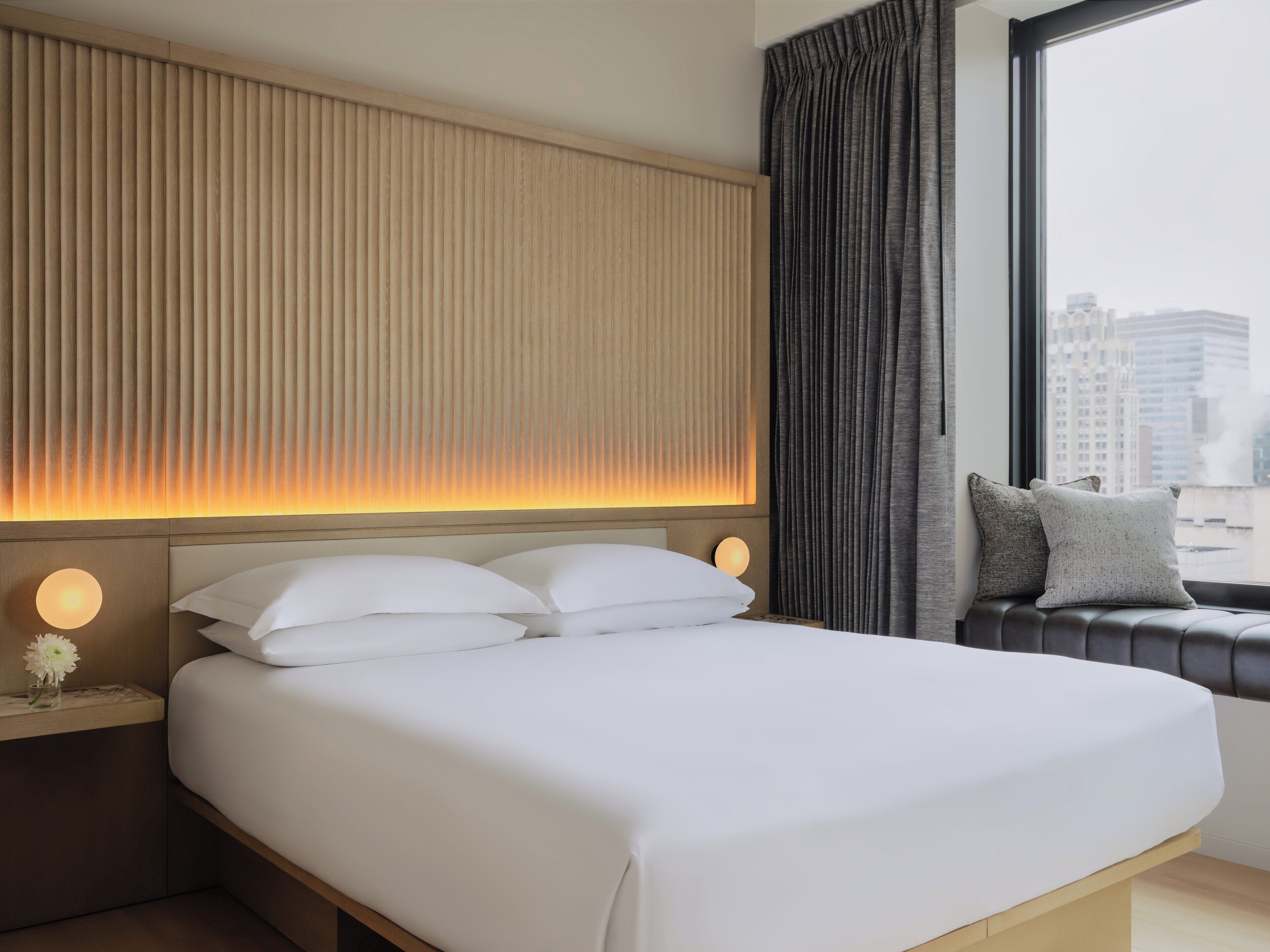 A hotel room with a large bed, a bedside table with a lamp, a window with a view of buildings, and a curtain.