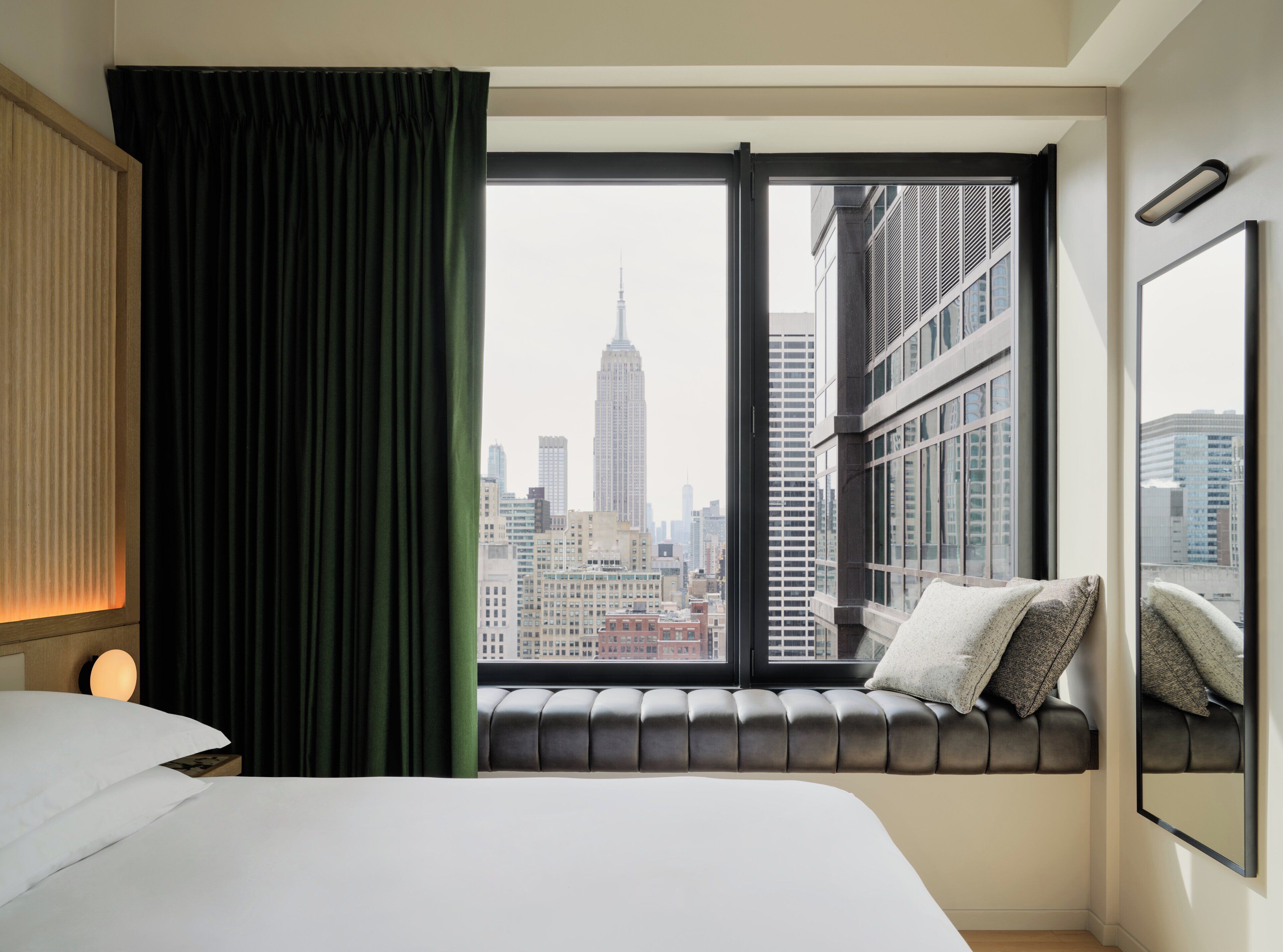 A hotel room with a large window, a bed, a headboard, a nightstand, and a wall-mounted lamp.
