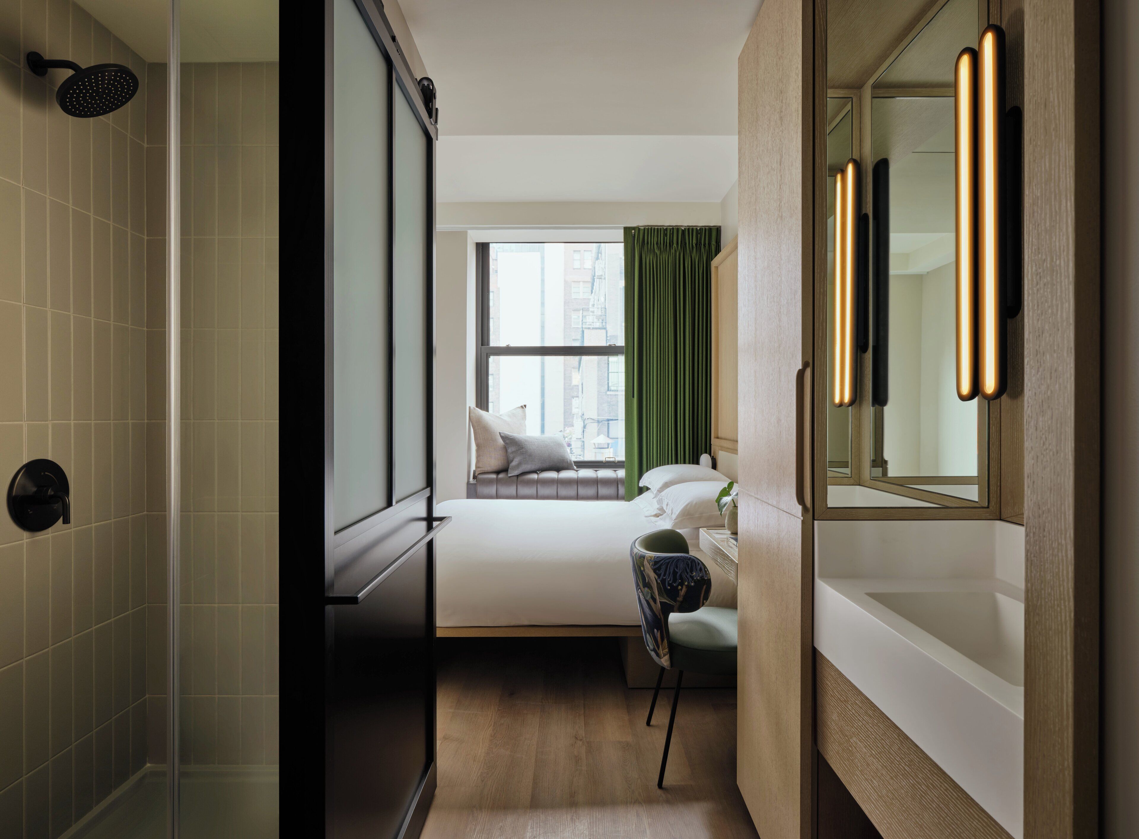 A modern hotel room with a shower, a bed with pillows, a chair, a window with curtains, and a mirror with lights.