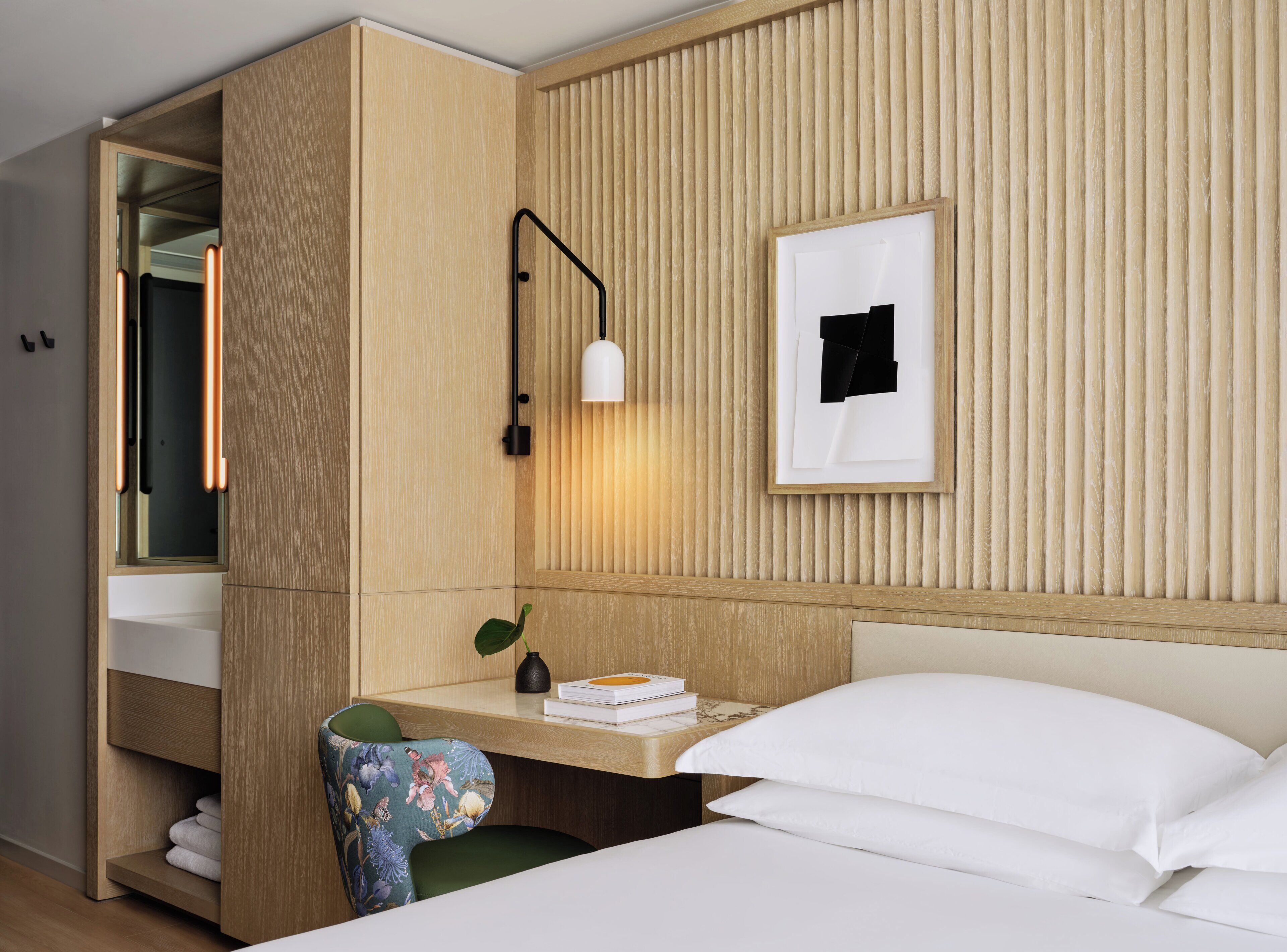 Une chambre d’hôtel comprenant un lit, un bureau avec une chaise, un miroir, un tableau encadré au mur et une table de chevet avec une lampe.