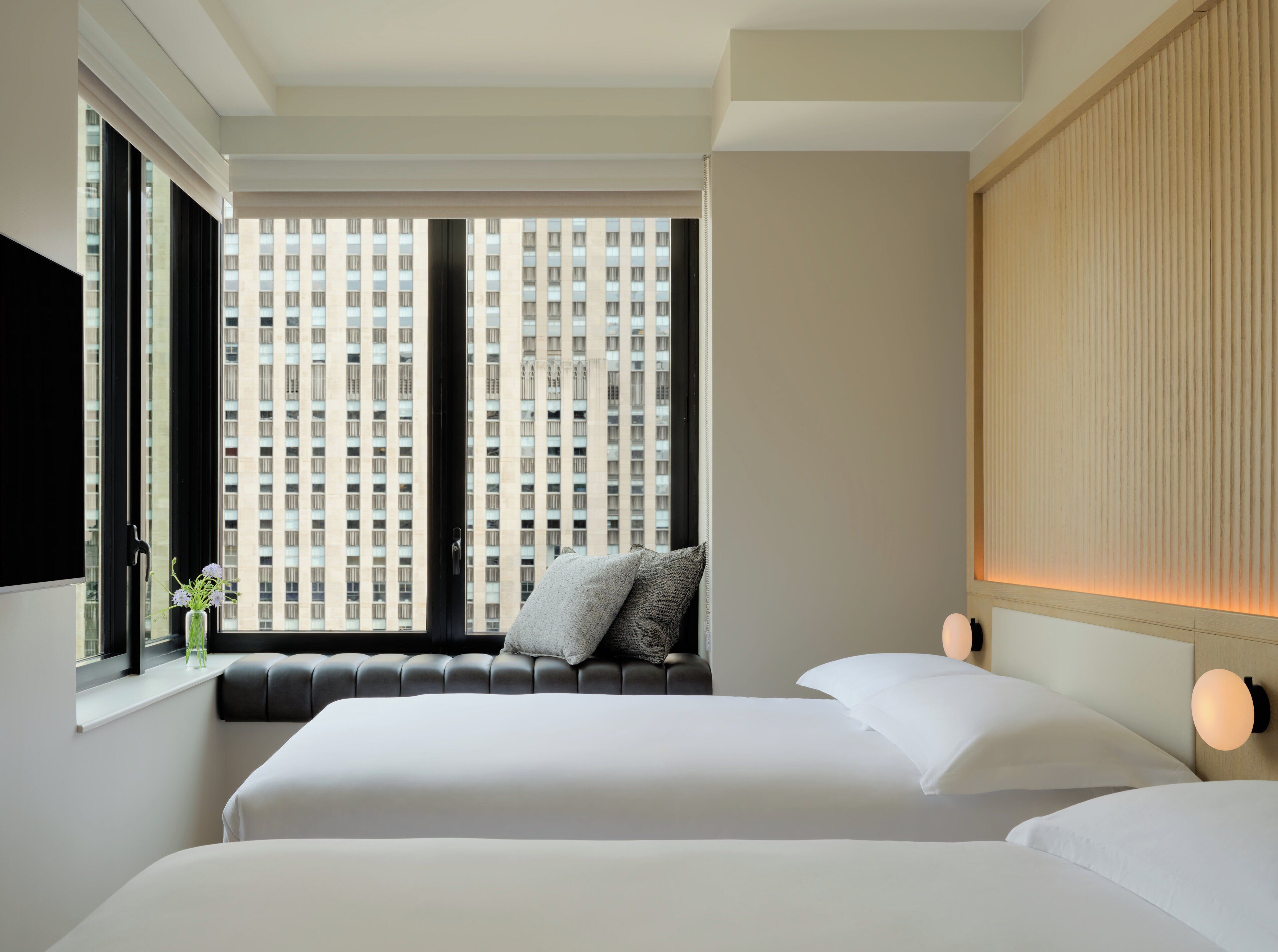 A hotel room with a large window, a bed, a seating area, and a wall-mounted light fixture.