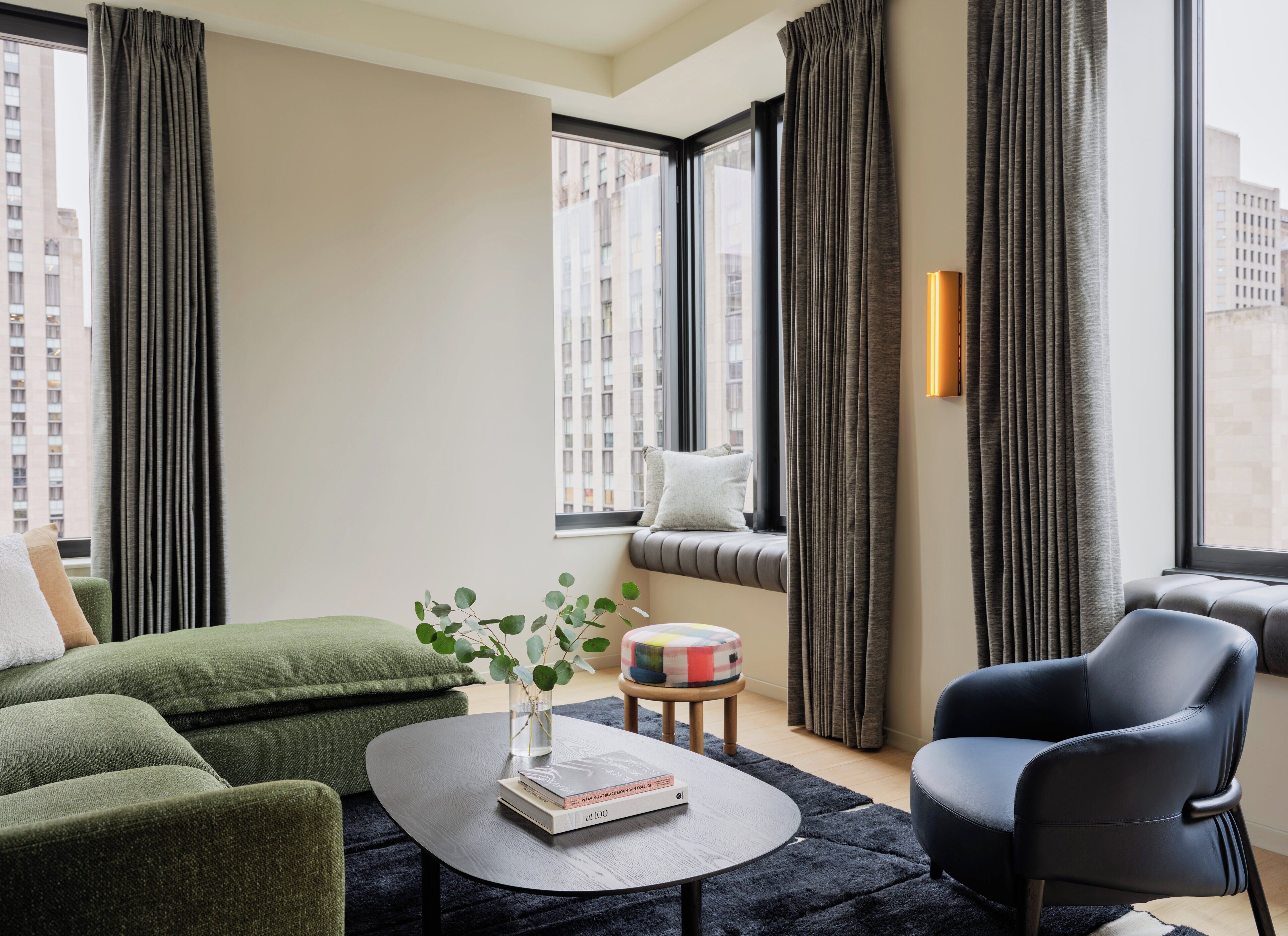 A modern living room with a green sofa, a black leather chair, a round coffee table with a book and a vase, and large windows with curtains.