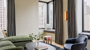 A modern living room with a green sofa, a black leather chair, a round coffee table with a book and a vase, and large windows with curtains.
