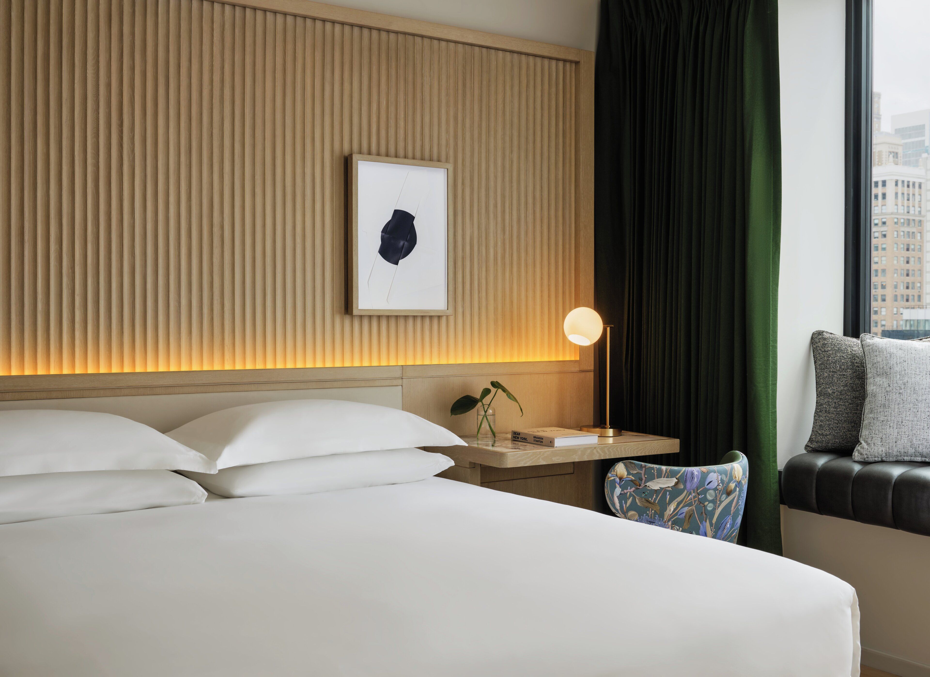 A hotel room with a bed, bedside table, lamp, artwork, and a view of the city through a window.