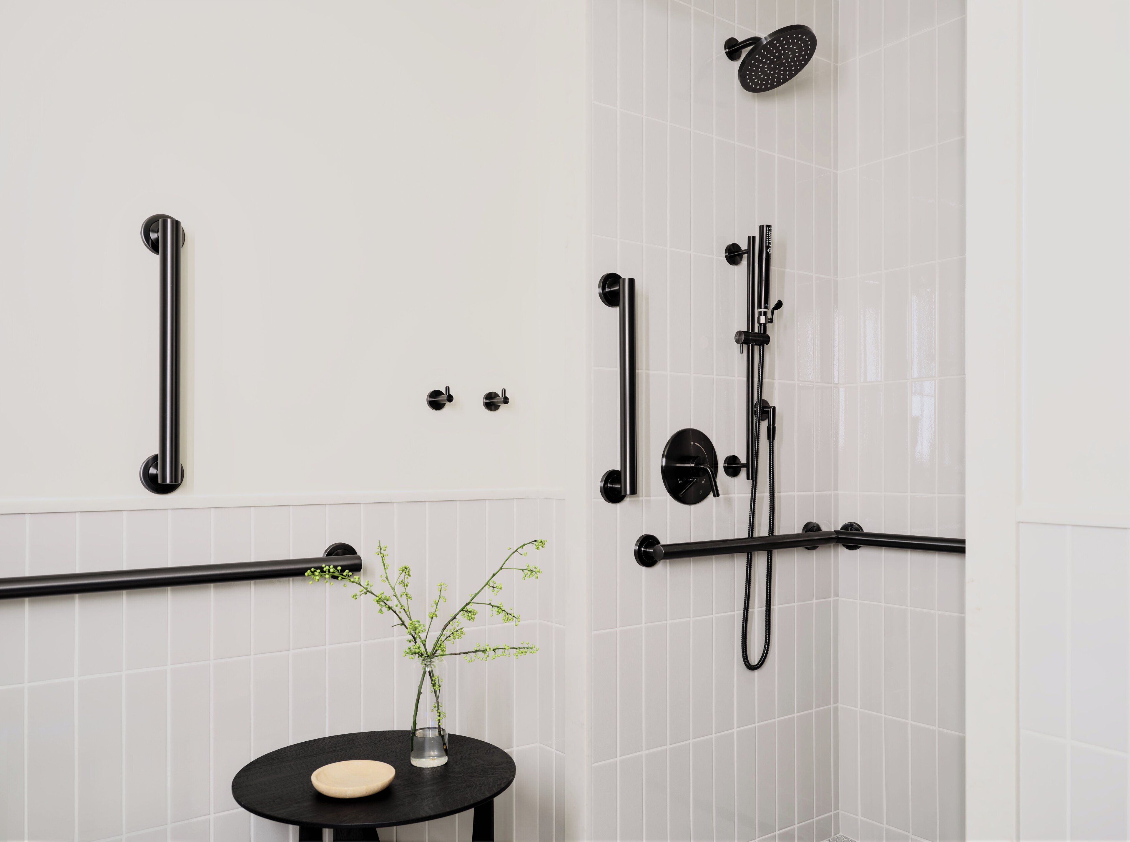 A bathroom with a shower, grab bars, a small table with a vase, and a soap dish.