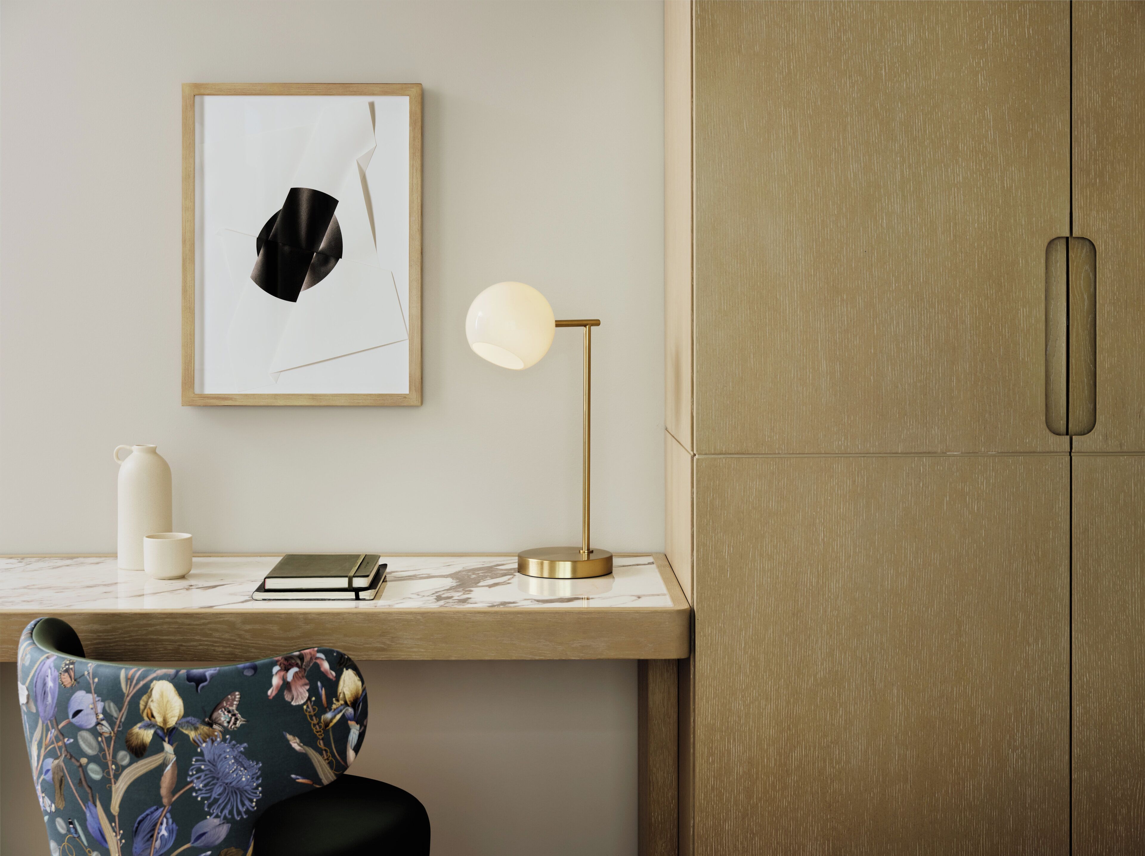 A desk with a framed artwork, a lamp, a notebook, and a decorative chair.