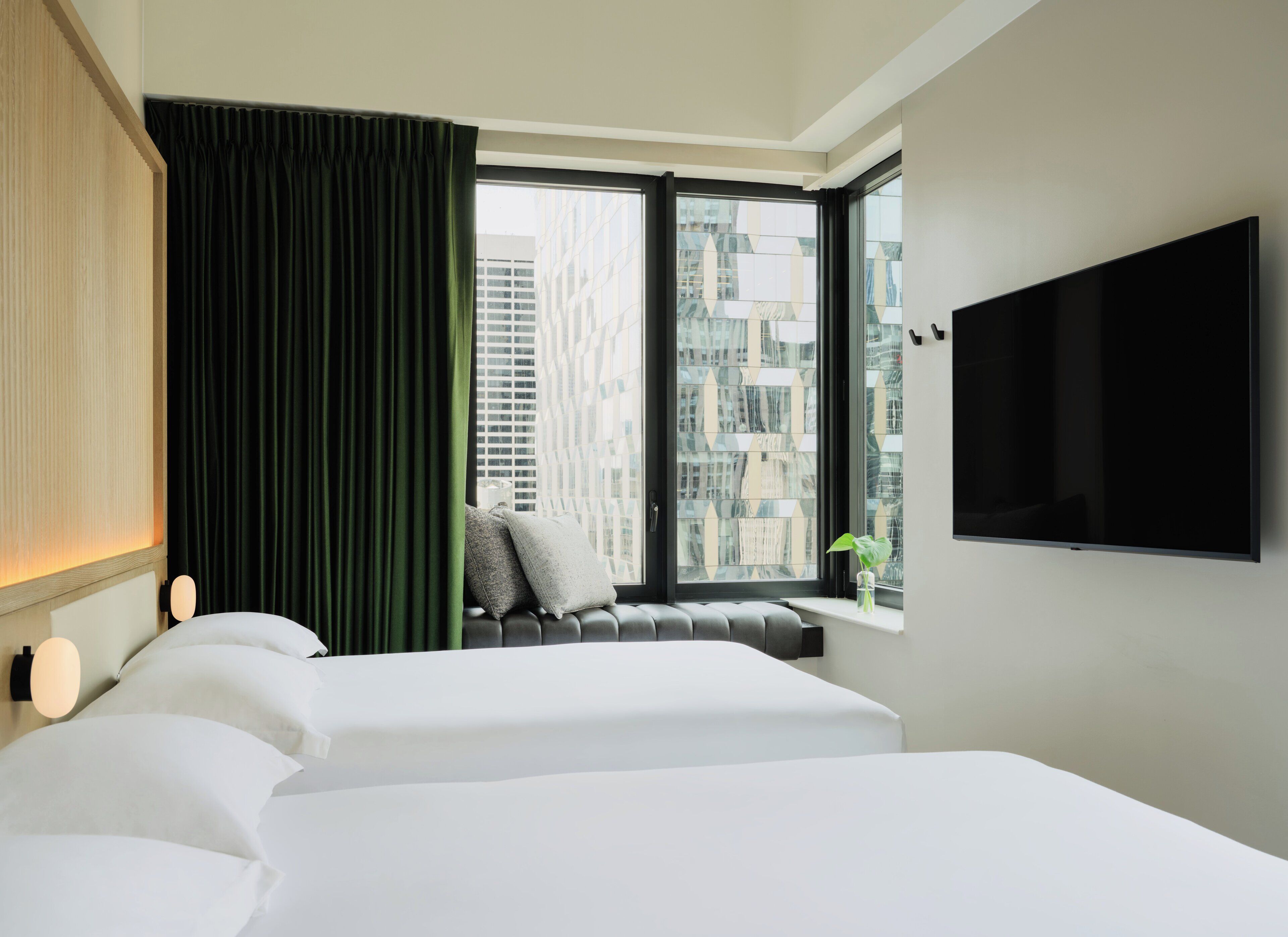 A hotel room with two beds, a large window with a view of buildings, a flat-screen TV mounted on the wall, and a green curtain.