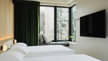A hotel room with two beds, a large window with a view of buildings, a flat-screen TV mounted on the wall, and a green curtain.