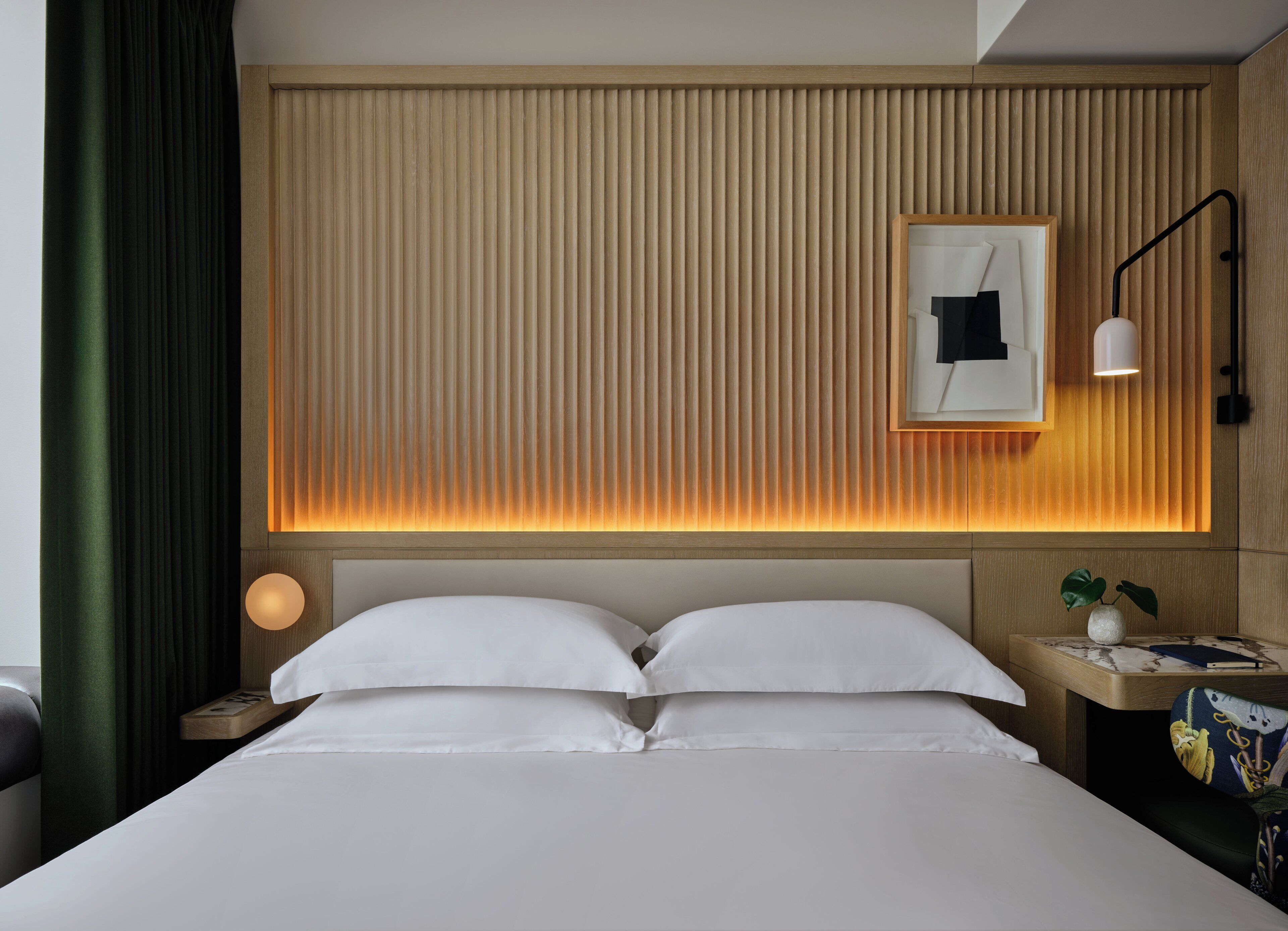 Una habitación de hotel con una cama, una mesita de noche con una lámpara, una mesita auxiliar y un estante en la pared con una planta.