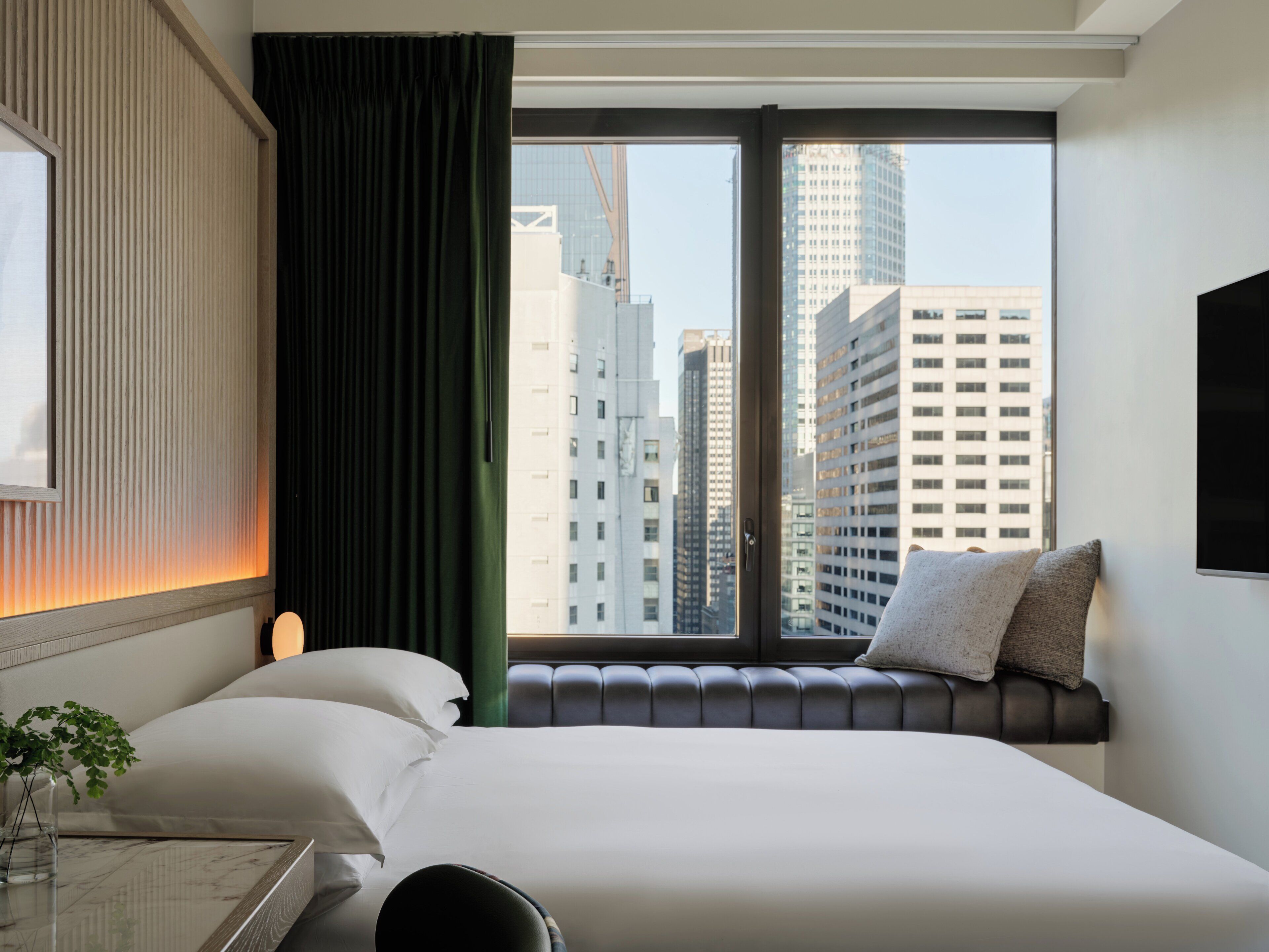 A hotel room with a large bed, a view of tall buildings, a window with a curtain, and a bedside table with a vase.