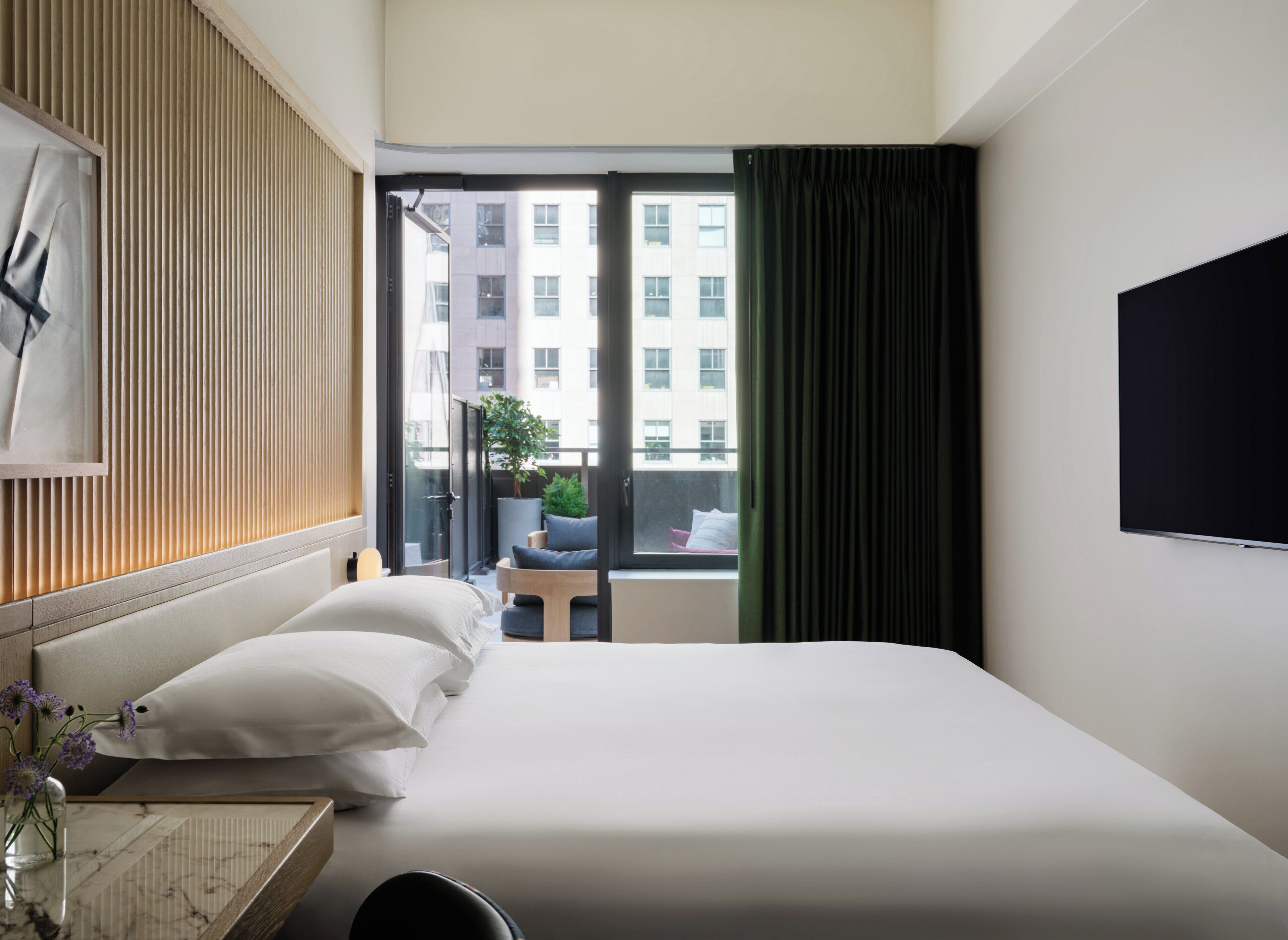 Une chambre d’hôtel avec un lit, un bureau agrémenté d’un vase de fleurs, une fenêtre avec des rideaux et une vue sur les bâtiments environnants.