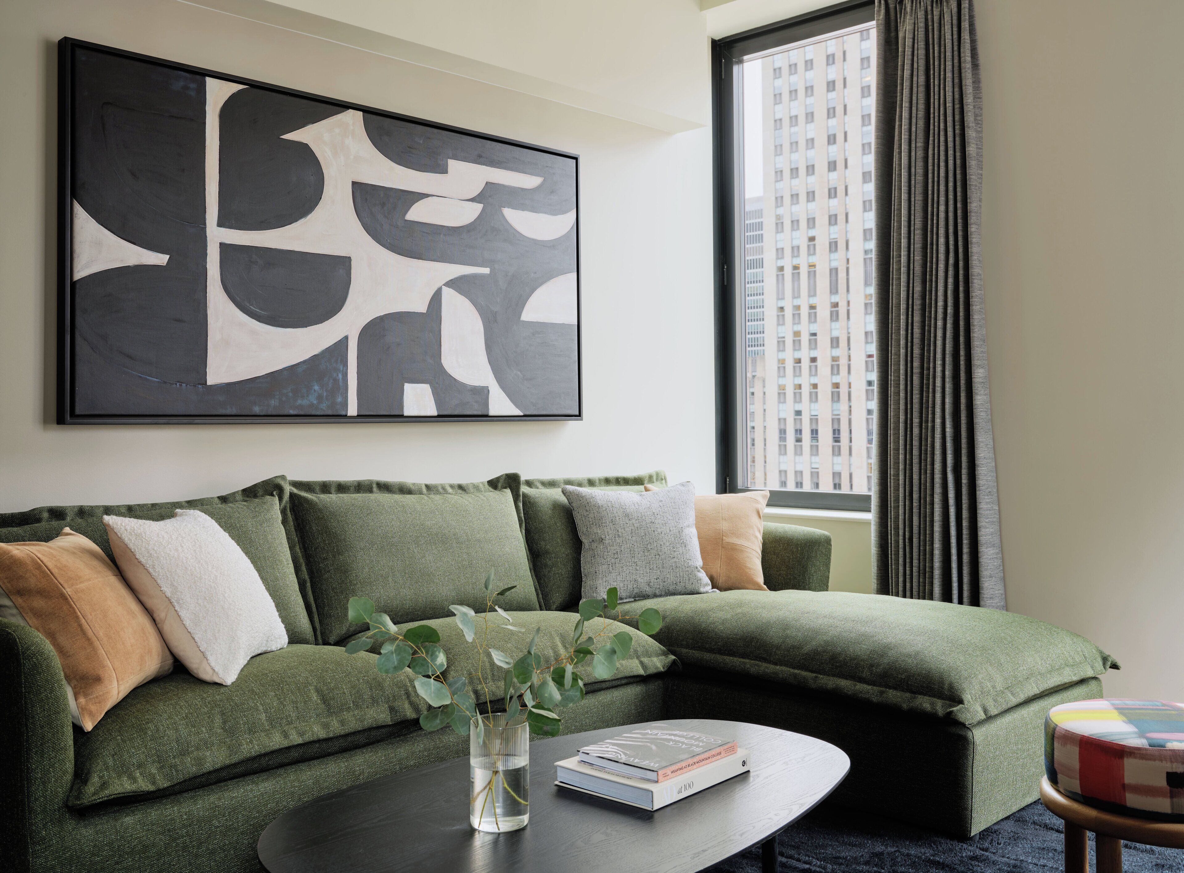 A modern living room with a green sectional sofa, a coffee table, a framed abstract painting, and a view of a cityscape through a window.