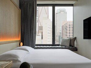 Une chambre d’hôtel avec une grande fenêtre, un lit, une banquette avec des coussins et un téléviseur fixé au mur.