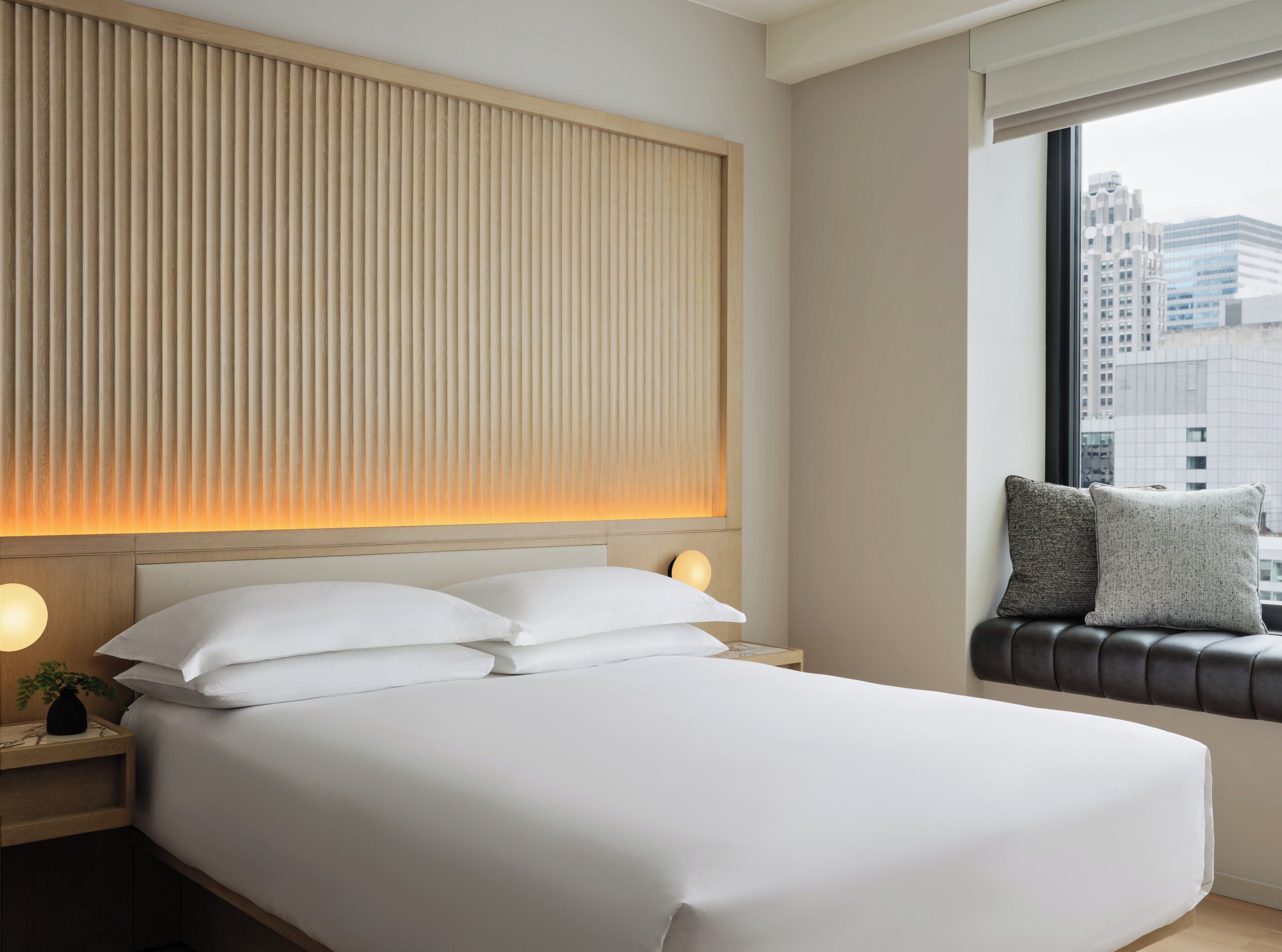 A hotel room with a large bed, a headboard with vertical slats, a bedside table with a lamp, and a view of the city through a window.