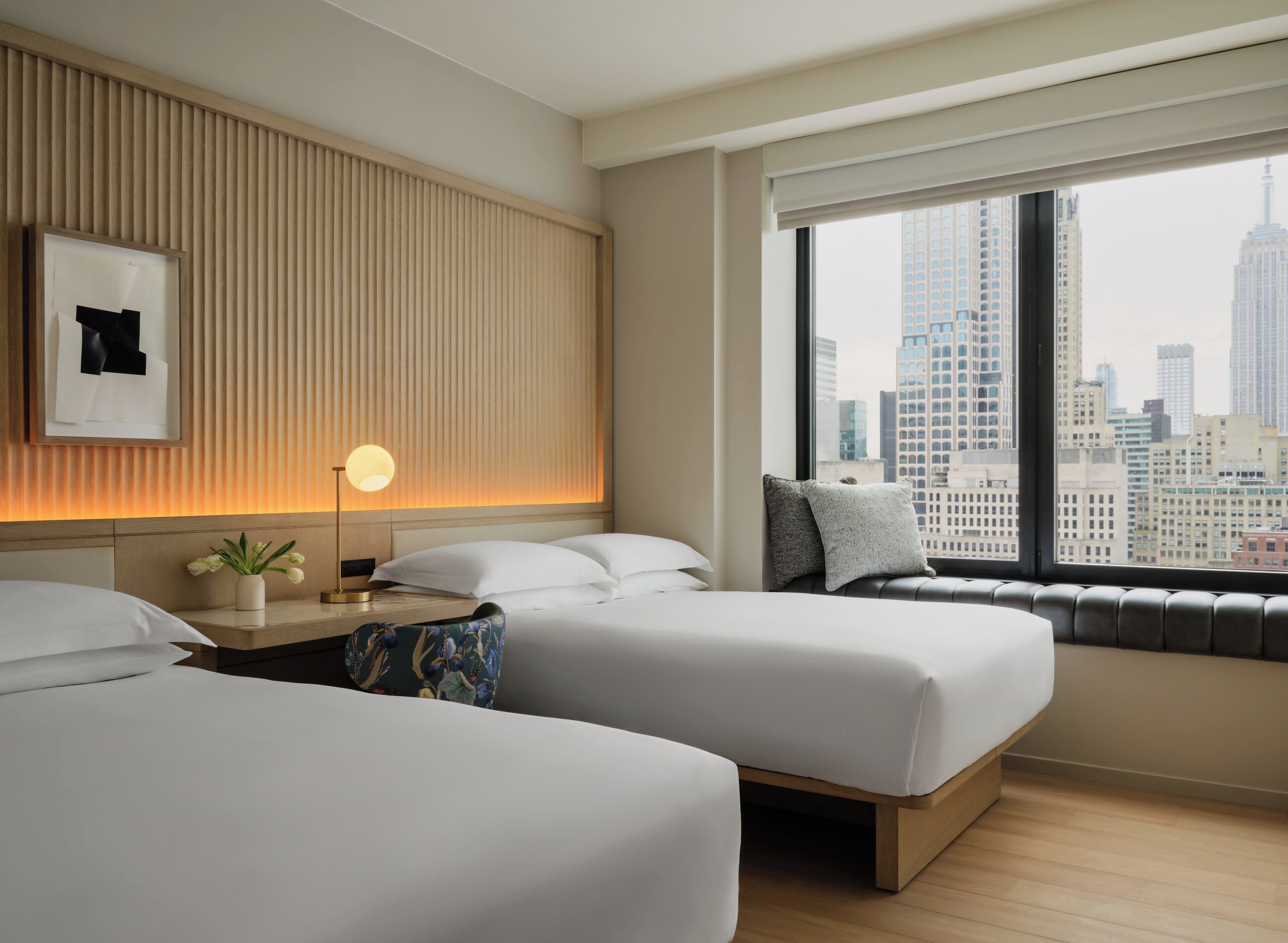 Habitación de hotel con dos camas, una mesita de noche con lámpara, una ventana con vistas a la ciudad y un cuadro colgado en la pared.