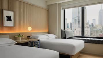 Habitación de hotel con dos camas, una mesita de noche con lámpara, una ventana con vistas a la ciudad y un cuadro colgado en la pared.