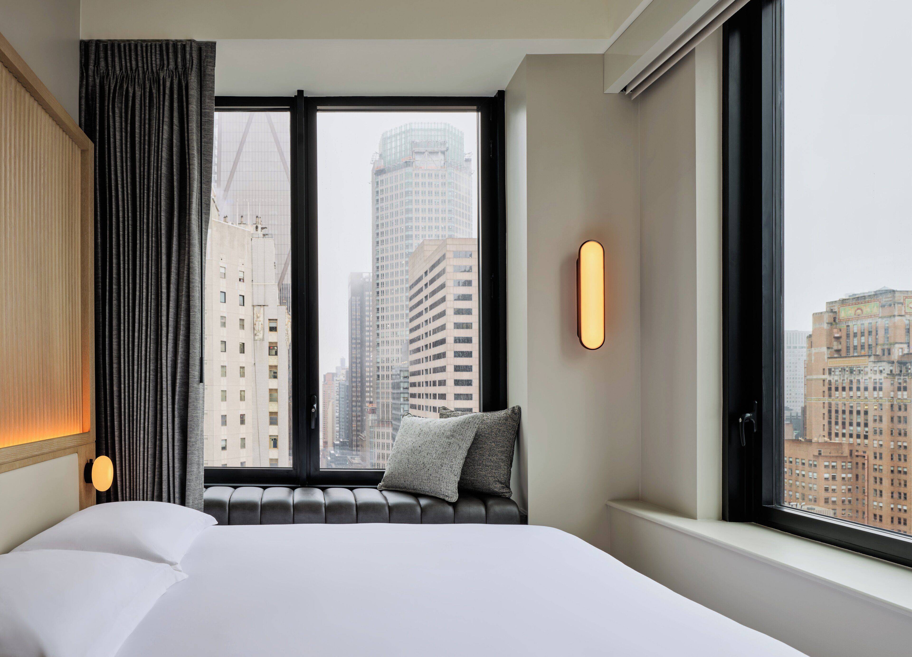 Quarto de hotel com uma cama grande, janela com vista para prédios altos, luminária na parede e cortina.