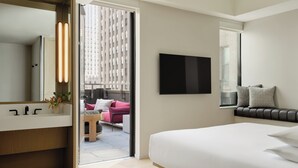 Una habitación de hotel moderna con una cama grande, un televisor de pantalla plana, una mesita de noche, un baño con espejo y vistas a la ciudad a través de un ventanal amplio.