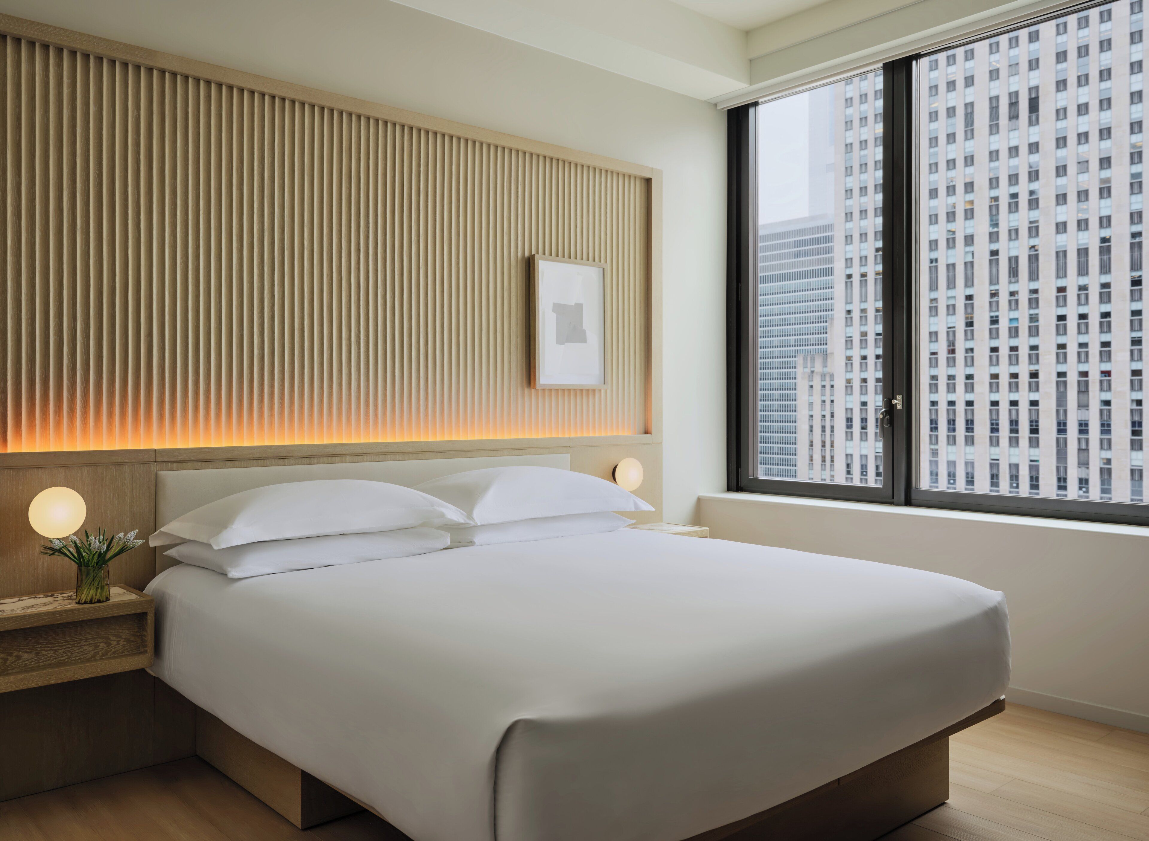 Une chambre d’hôtel moderne avec un grand lit, une table de chevet avec une lampe, un tableau accroché au mur et une grande fenêtre donnant sur la ville.