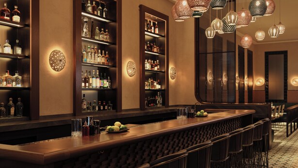 Una zona de bar con estantes de botellas de licor, luces decorativas colgantes y una barra larga con taburetes.