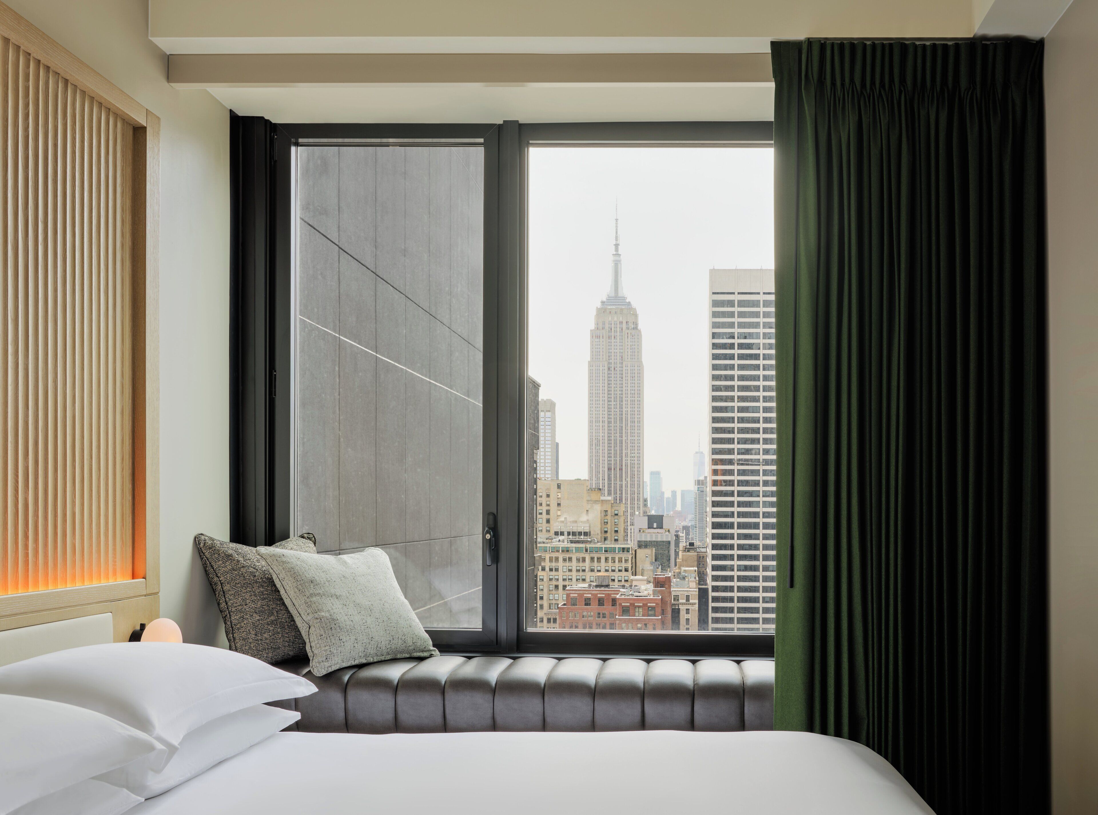 A hotel room with a large window, a view of the Empire State Building, a bed with pillows, and a green curtain.