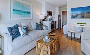 A modern living room with a sofa, coffee table, and a kitchen in the background. - 1 Bedroom Oceanfront - Walk right out to the beach! (Ocean Isle Beach)