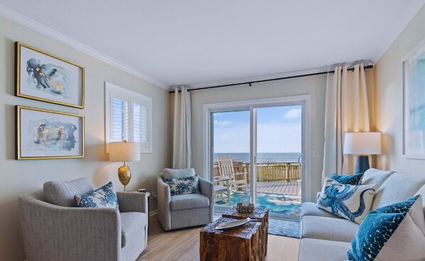 A living room with a view of the ocean, a balcony with a wooden deck, and two framed artworks on the wall. - 1 Bedroom Oceanfront - Walk right out to the beach! (Ocean Isle Beach)