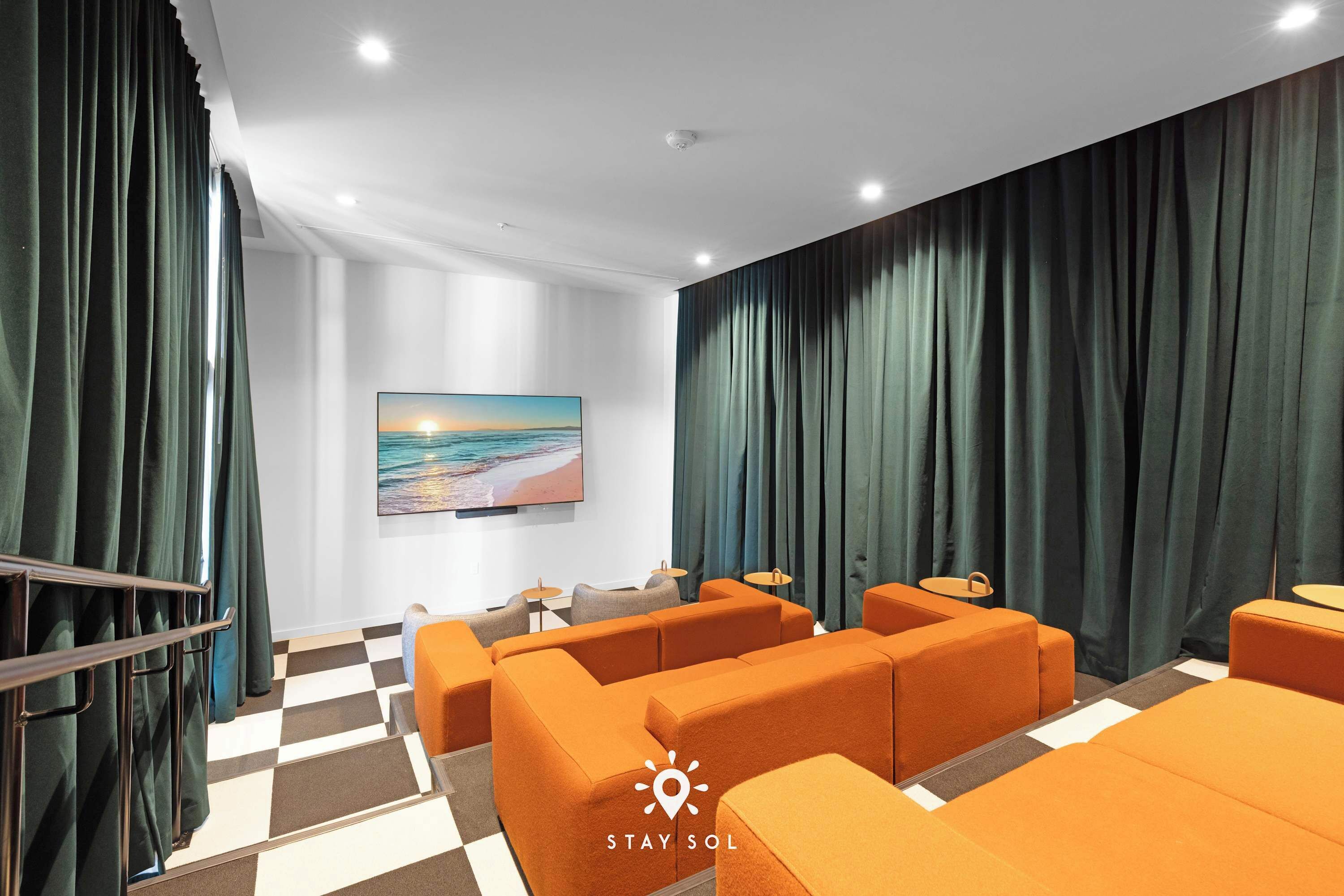 A hotel room with orange seating, a checkered floor, a painting of a sunset, and dark curtains.