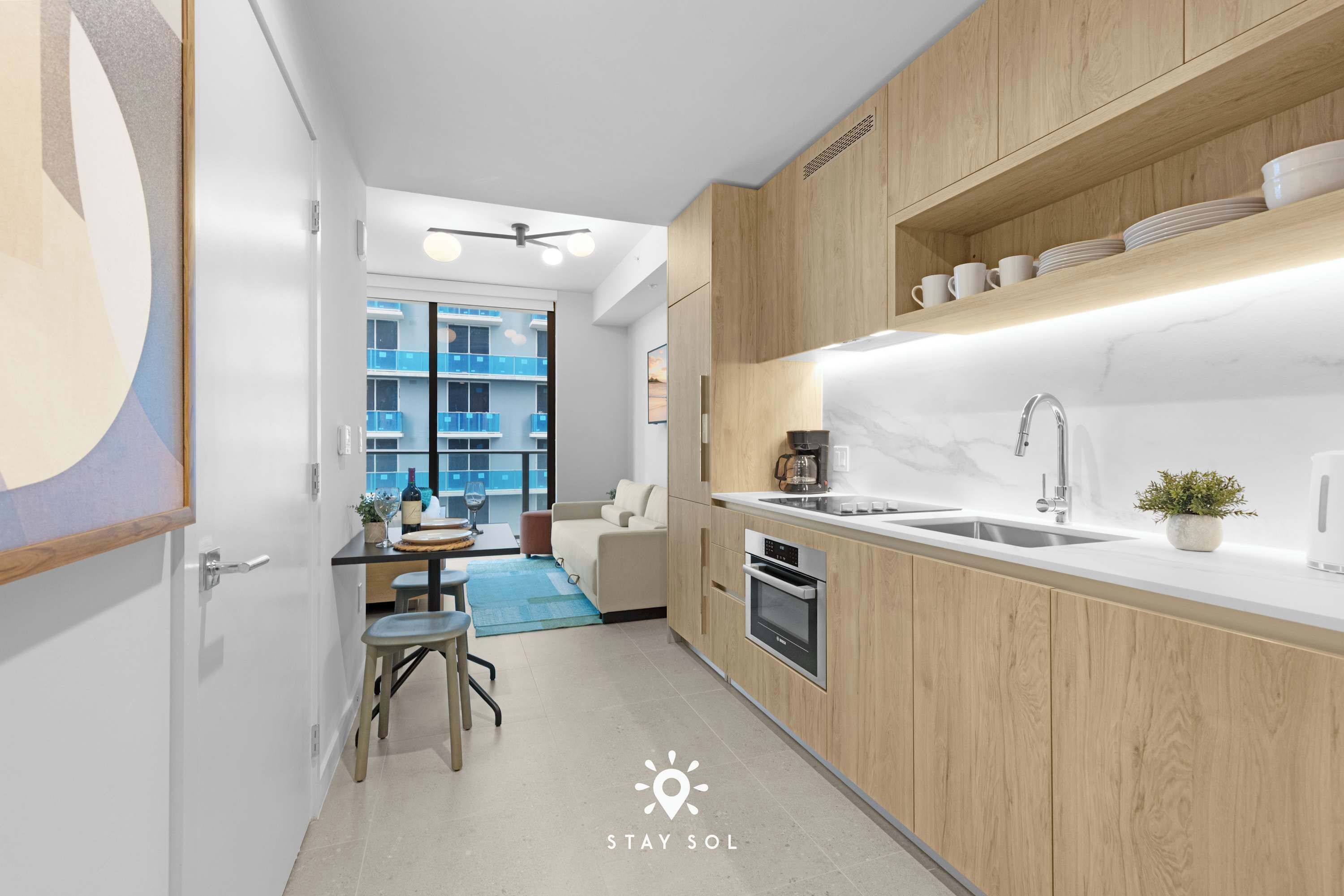 A modern kitchen with wooden cabinets, a sink, and a countertop. There is a dining area with a table and chairs, and a view of a blue building outside.