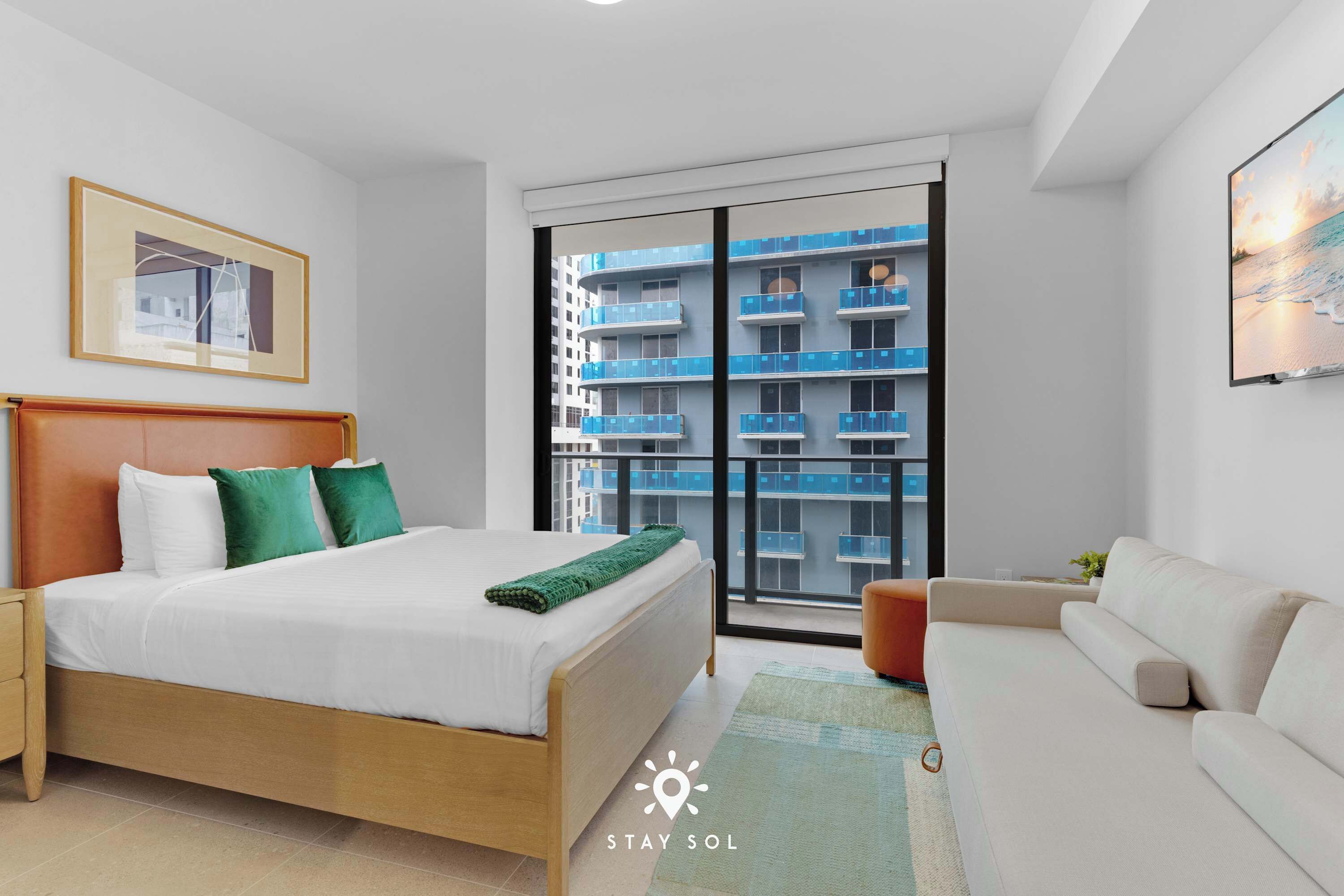 Ett modernt hotellrum med en säng, en soffa, ett stort fönster med utsikt över en byggnad och en inramad tavla på väggen.