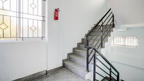Uma escada com corrimão de metal, um extintor fixado na parede e uma janela com padrão geométrico.
