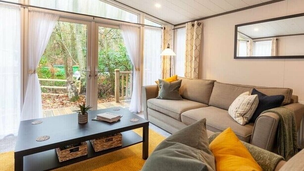 A living room with a sofa, cushions, a coffee table, and a large window with curtains. - Wildwood - Kelling Heath Holiday Park - 16335 (Holt)