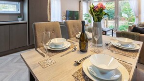 A dining area with a table set for four, including wine glasses, plates, and cutlery. There is a vase with flowers on the table, and the room has a view of greenery outside. - Wildwood - Kelling Heath Holiday Park - 16335 (Holt)