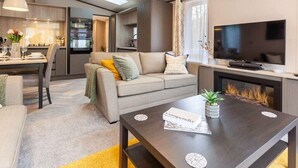 A modern living room with a sofa, coffee table, and TV. There is a kitchen area in the background with a dining table and chairs. - Wildwood - Kelling Heath Holiday Park - 16335 (Holt)
