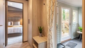 A hallway leading to a bedroom with a bed, a small table with a plant, and a door to a balcony with a view of the outdoors. - Wildwood - Kelling Heath Holiday Park - 16335 (Holt)