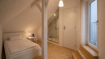 A bedroom with a bed, a nightstand, a small table, a shower, and a window.