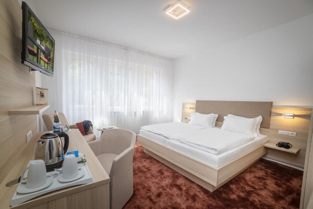 A hotel room with a bed, a desk with a kettle, a chair, and a TV on the wall.
