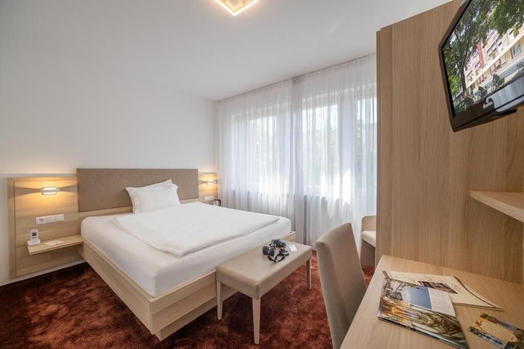 A hotel room with a bed, bedside table, chair, desk, and a TV mounted on the wall.