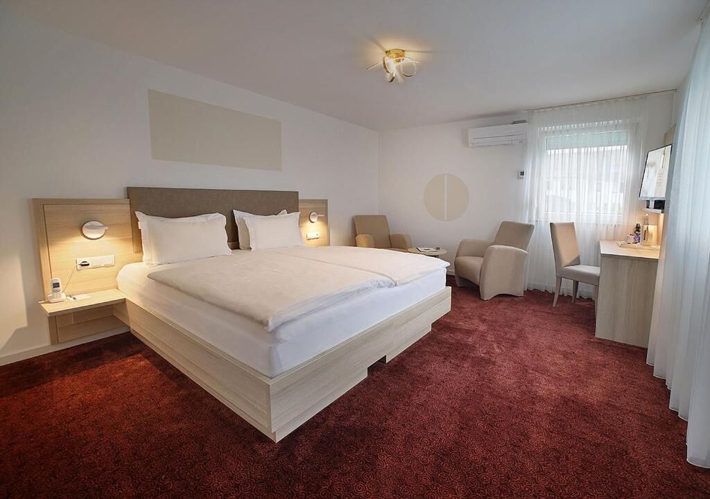 Een hotelkamer met een groot bed, twee nachtkastjes met lampen, een bureau met een stoel en een groot raam met gordijnen.