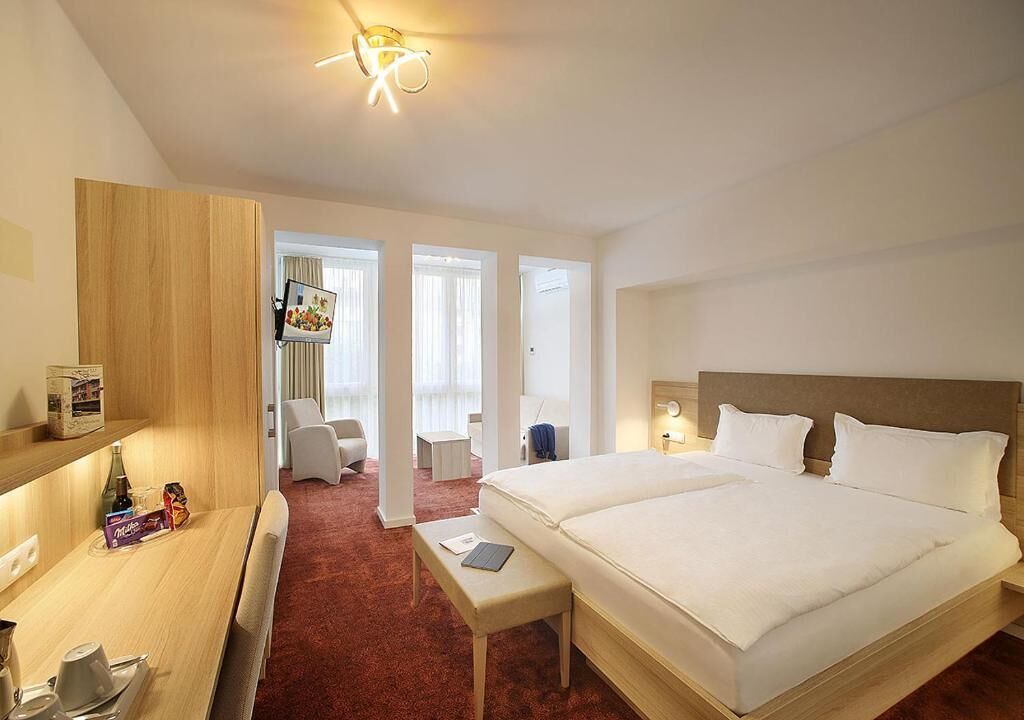 Habitación de hotel con una cama grande, un escritorio con silla, una mesita pequeña y un armario. Se ve una ventana con cortinas y un marco de fotos en la pared.