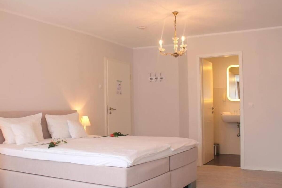 A bedroom with a large bed, bedside lamps, a chandelier, and a bathroom visible through an open door.