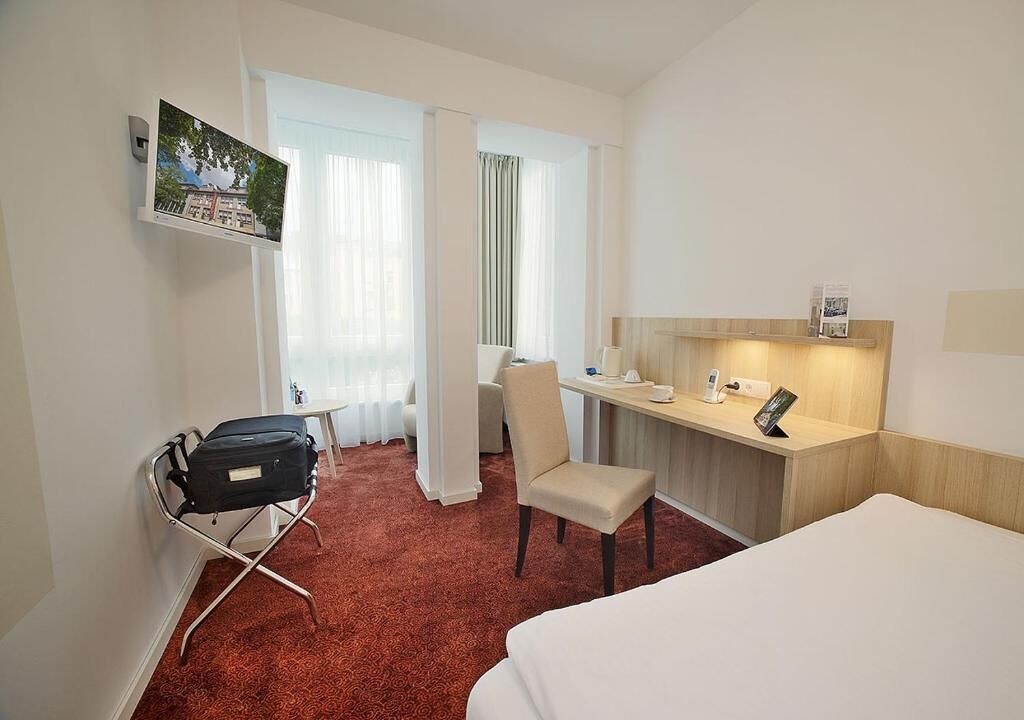 Ein Hotelzimmer mit einem Bett, einem Schreibtisch mit Stuhl, einem Koffer und einem an der Wand montierten Fernseher.