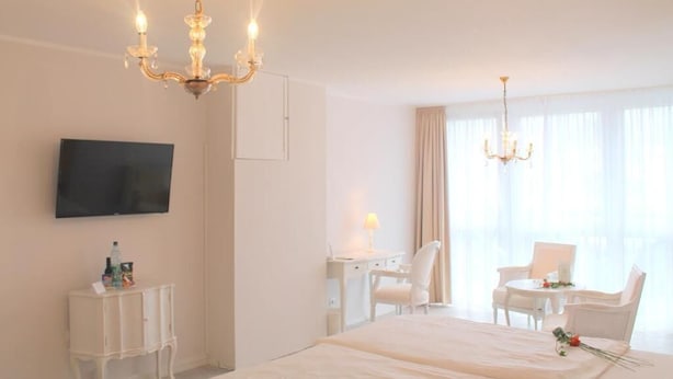 A hotel room with a large bed, a desk with a chair, a TV mounted on the wall, and a chandelier.