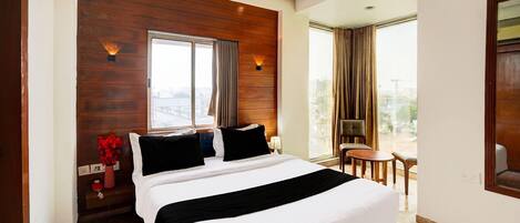 A hotel room with a large bed, wooden panel wall, two windows with curtains, a small table and chair, and a view of the city.