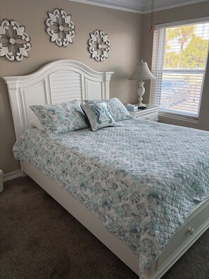 Un dormitorio con cama blanca, ropa de cama con estampado floral, decoración en la pared, una mesita de noche con lámpara y una ventana con persianas.